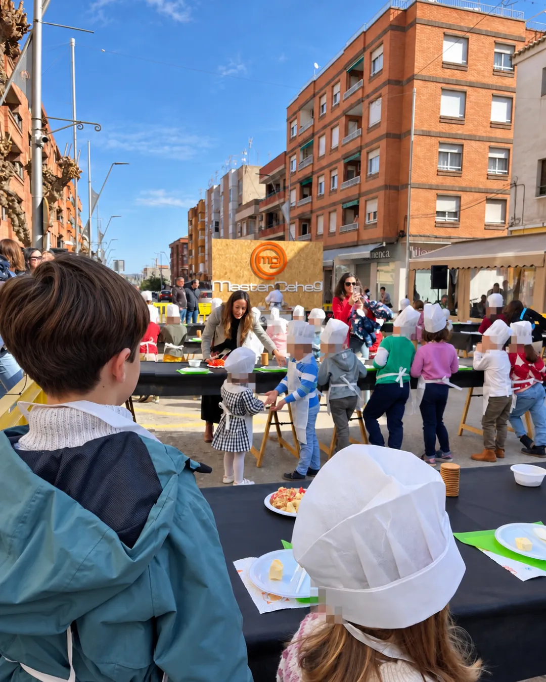 Niños con gorro y delantal de cocinero participan en un taller de cocina al aire libre de MasterCehegín, con mesas en la calle y familias alrededor.